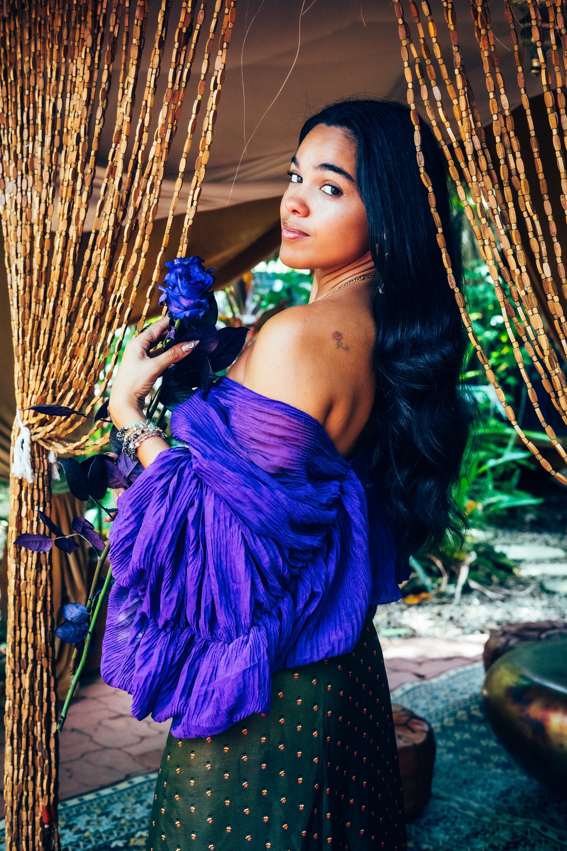Woman in a purple top and green skirt holding flowers, standing in front of decorative curtains.