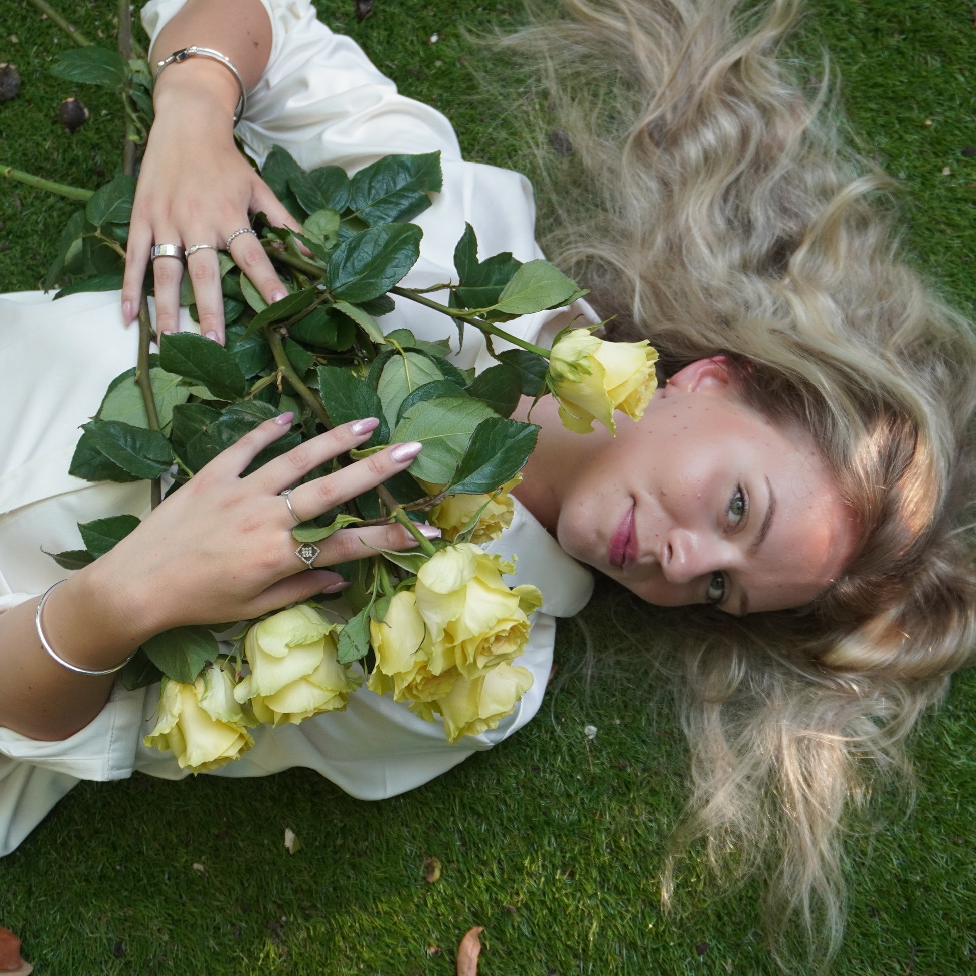 Woman lying on grass with yellow roses, wearing a white dress.