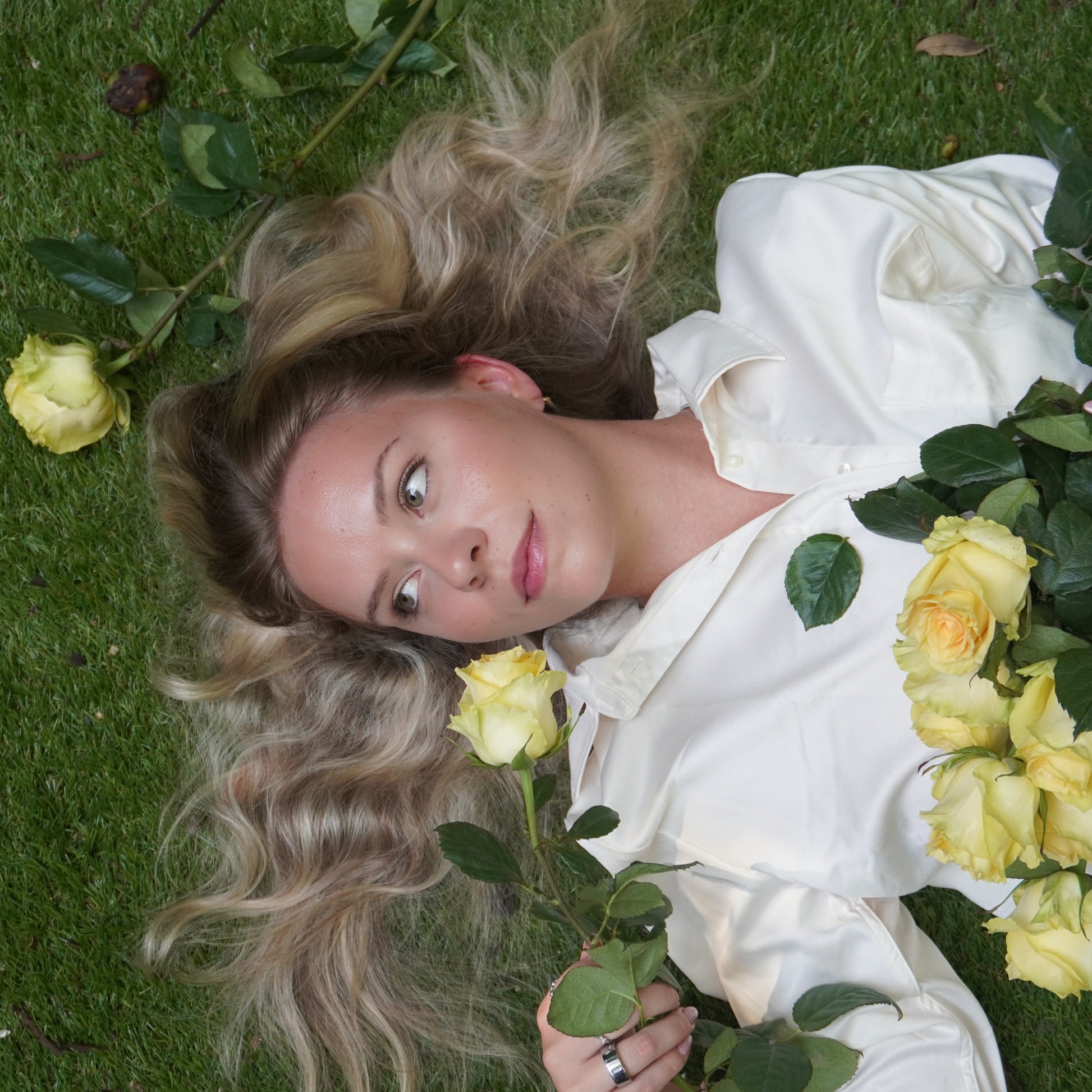 Woman lying on grass with yellow roses, wearing a white outfit