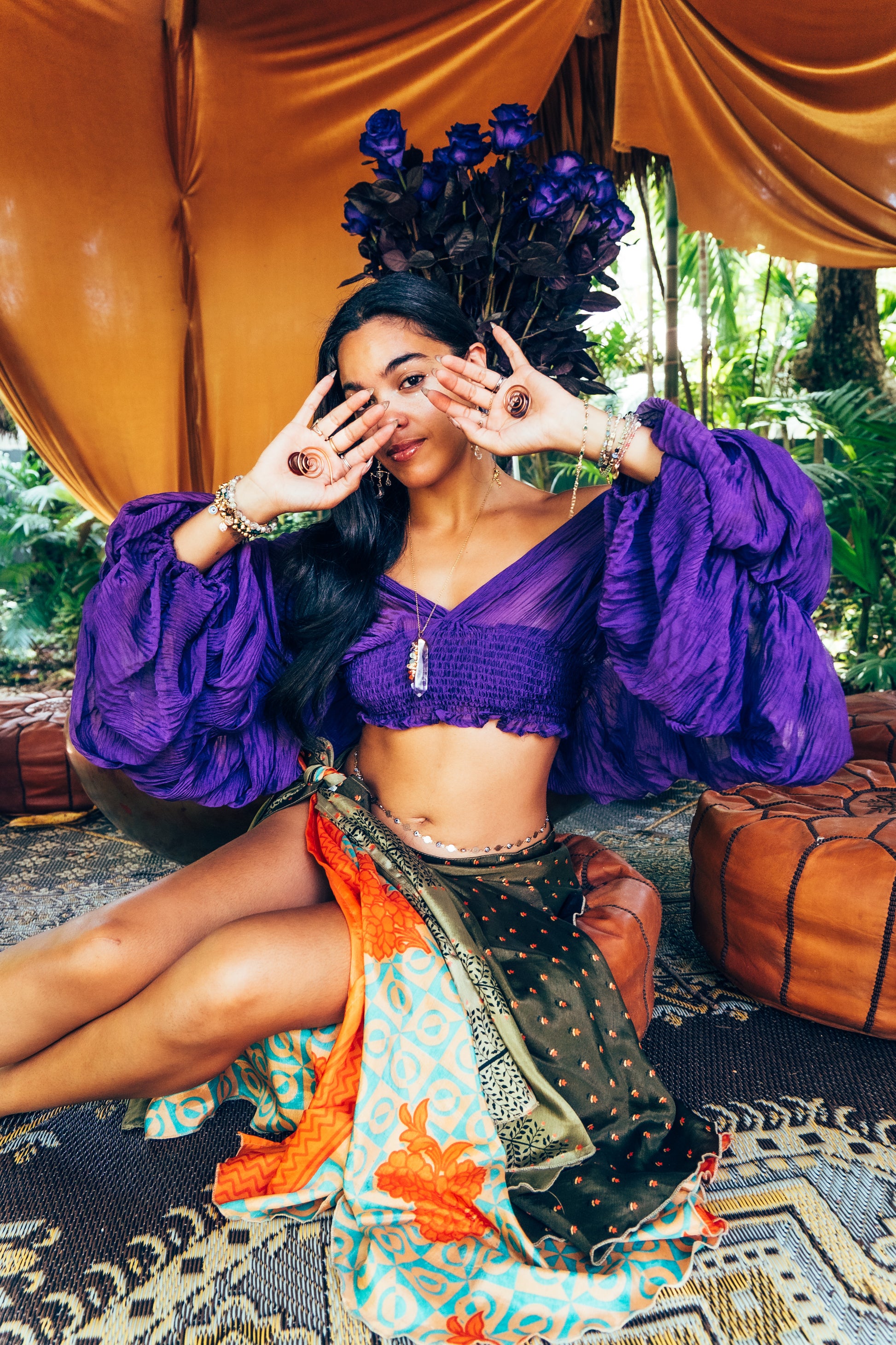 Woman in a vibrant outfit with a purple top and colorful skirt, sitting on a patterned rug with a floral arrangement.