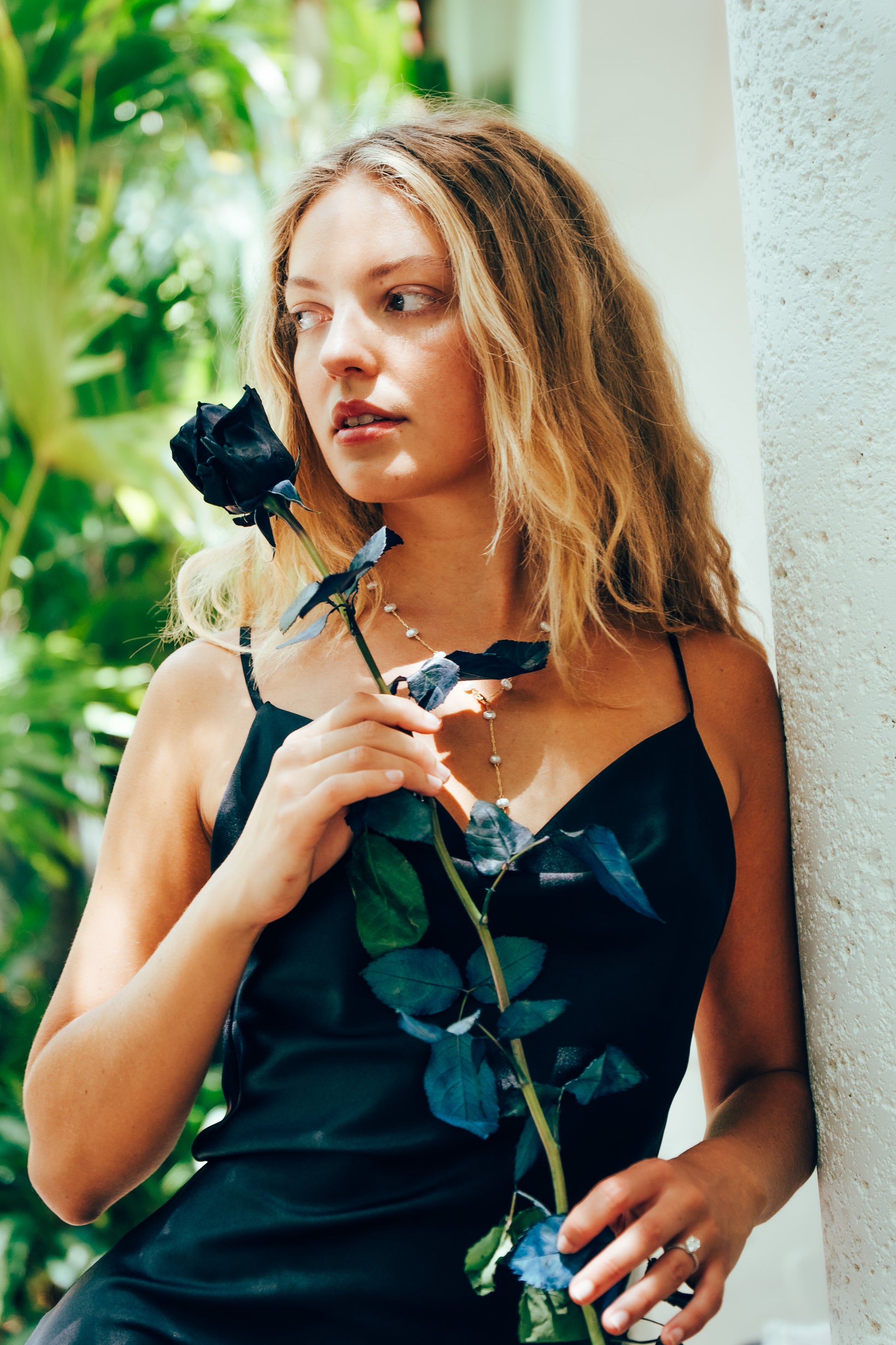 Woman holding a black rose against a white wall with greenery