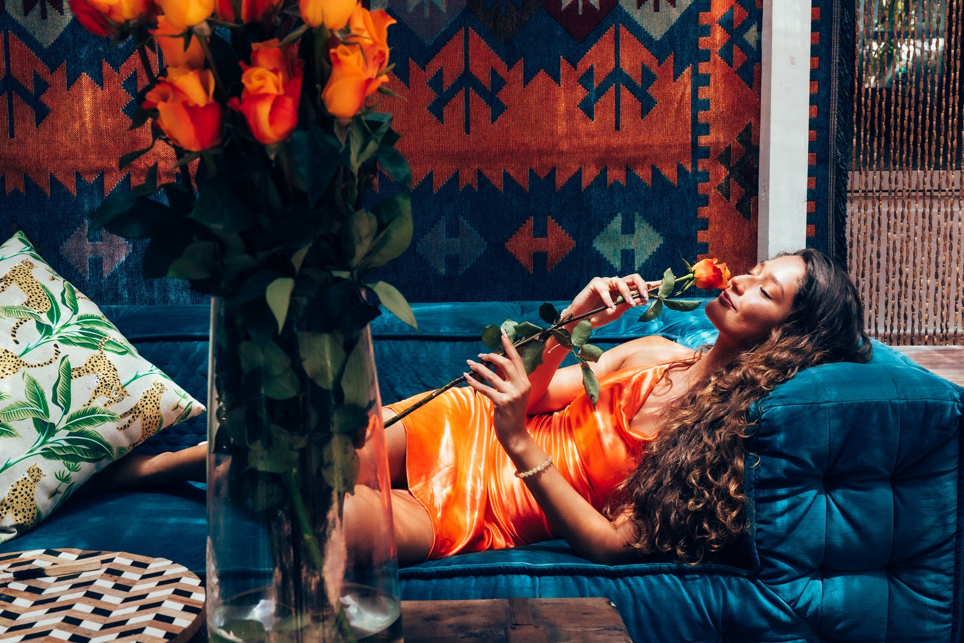 Woman lying on a blue couch holding a red rose, with a vase of flowers and patterned wall in the background.