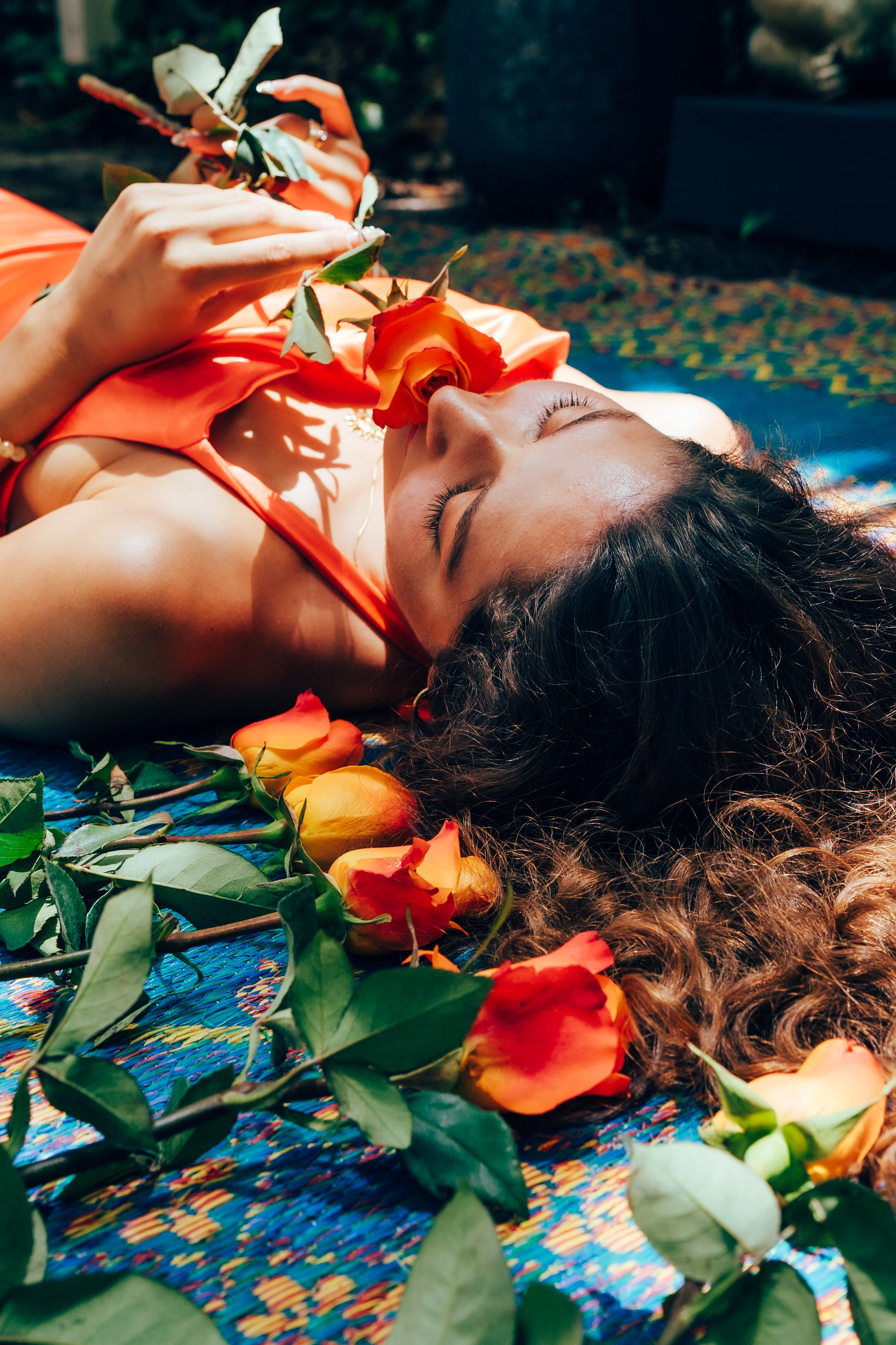 Woman lying on a colorful blanket with flowers around her, wearing an orange top.
