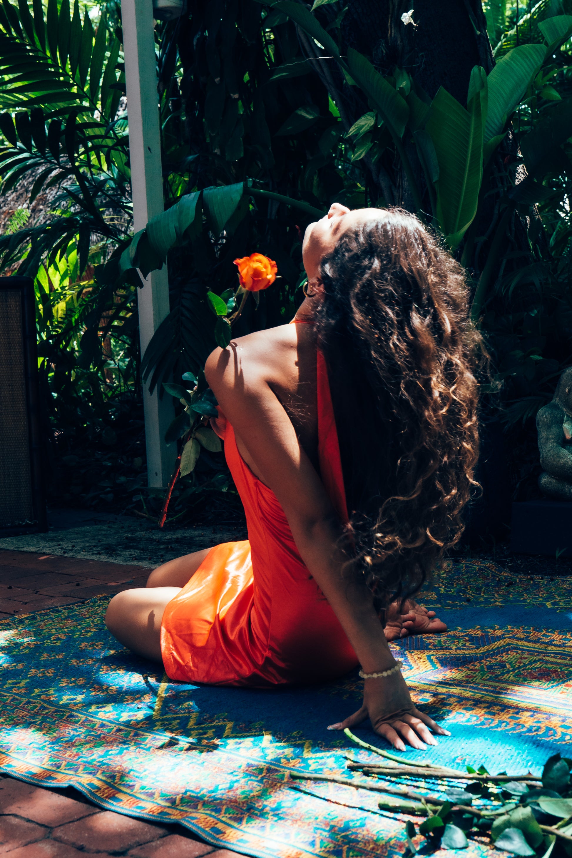Woman in an orange dress sitting on a colorful mat with a flower in a garden setting