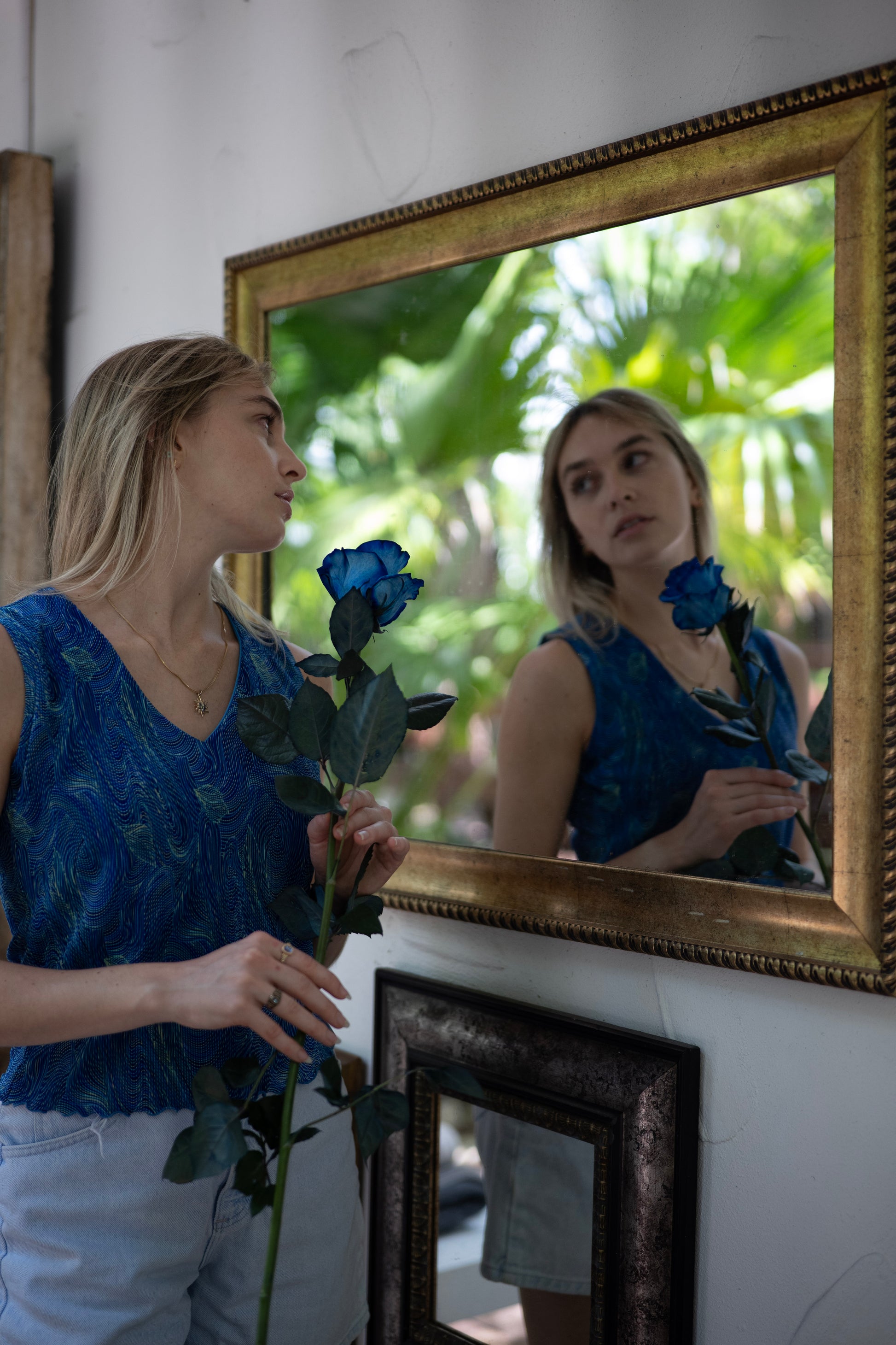 Woman holding a blue rose in front of a mirror with a gold frame.