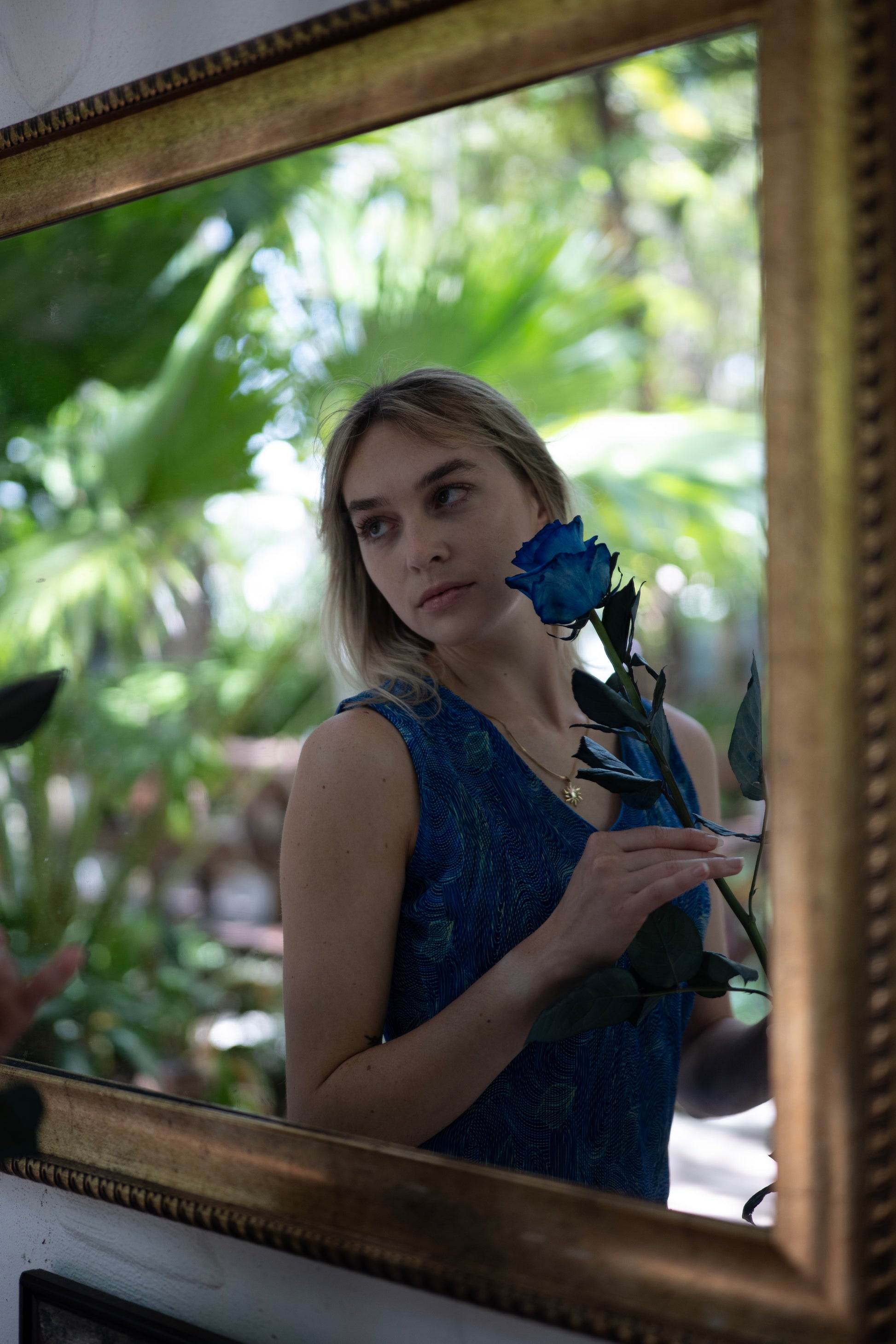 Woman holding a blue flower in front of a mirror with a gold frame.