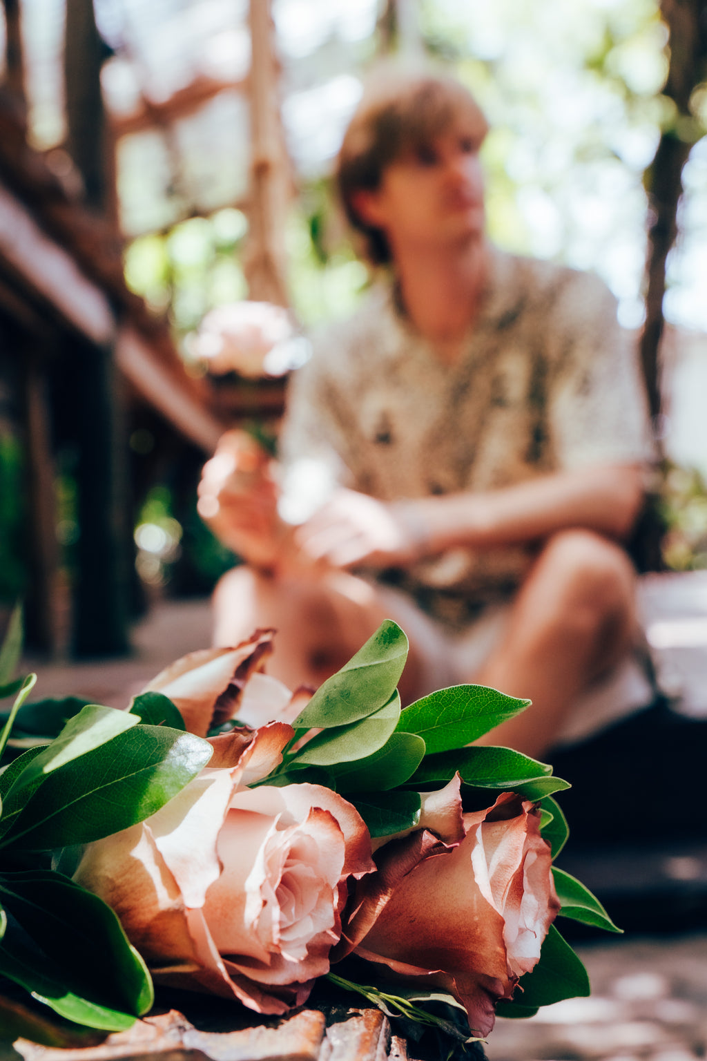 Bouquet of pink roses with green leaves in the foreground, blurred person sitting outdoors in the background.
